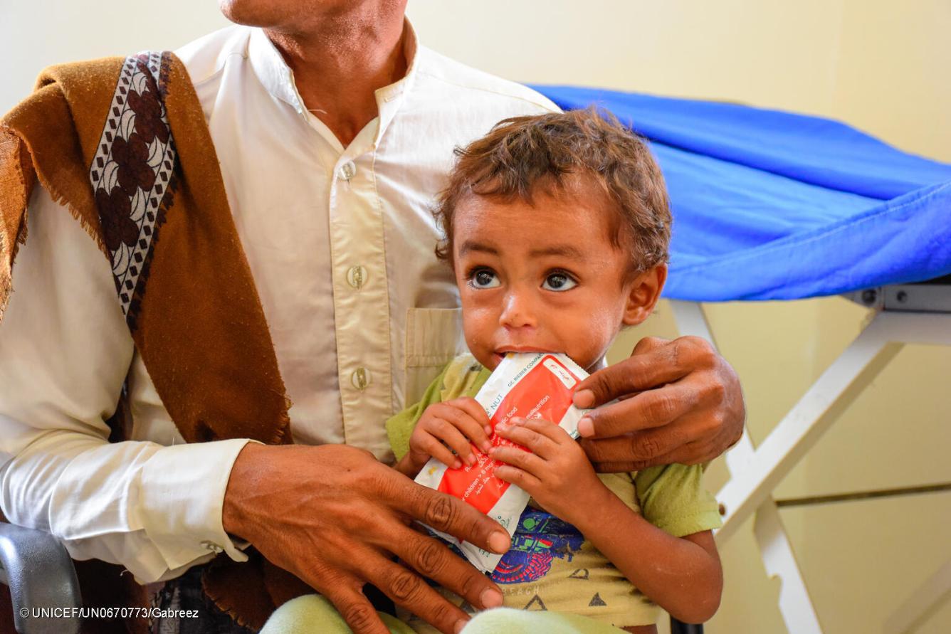 Un jeune garçon assis sur les genoux d’un homme mange un aliment thérapeutique en sachet dans un centre de traitement de la malnutrition au Yémen.
