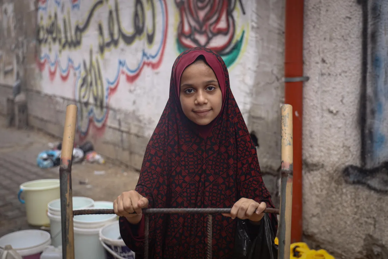 A girl stands near some water taps, she is looking directly at the camera.
