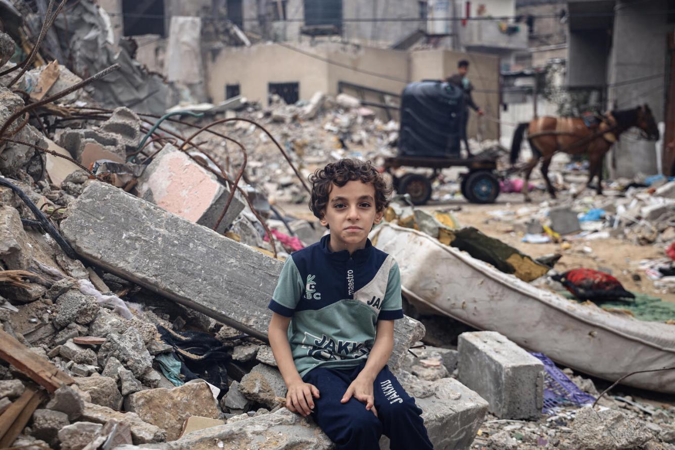 Un enfant assis sur les décombres de la maison familiale regarde l’objectif. On voit derrière lui tous les débris et gravats.