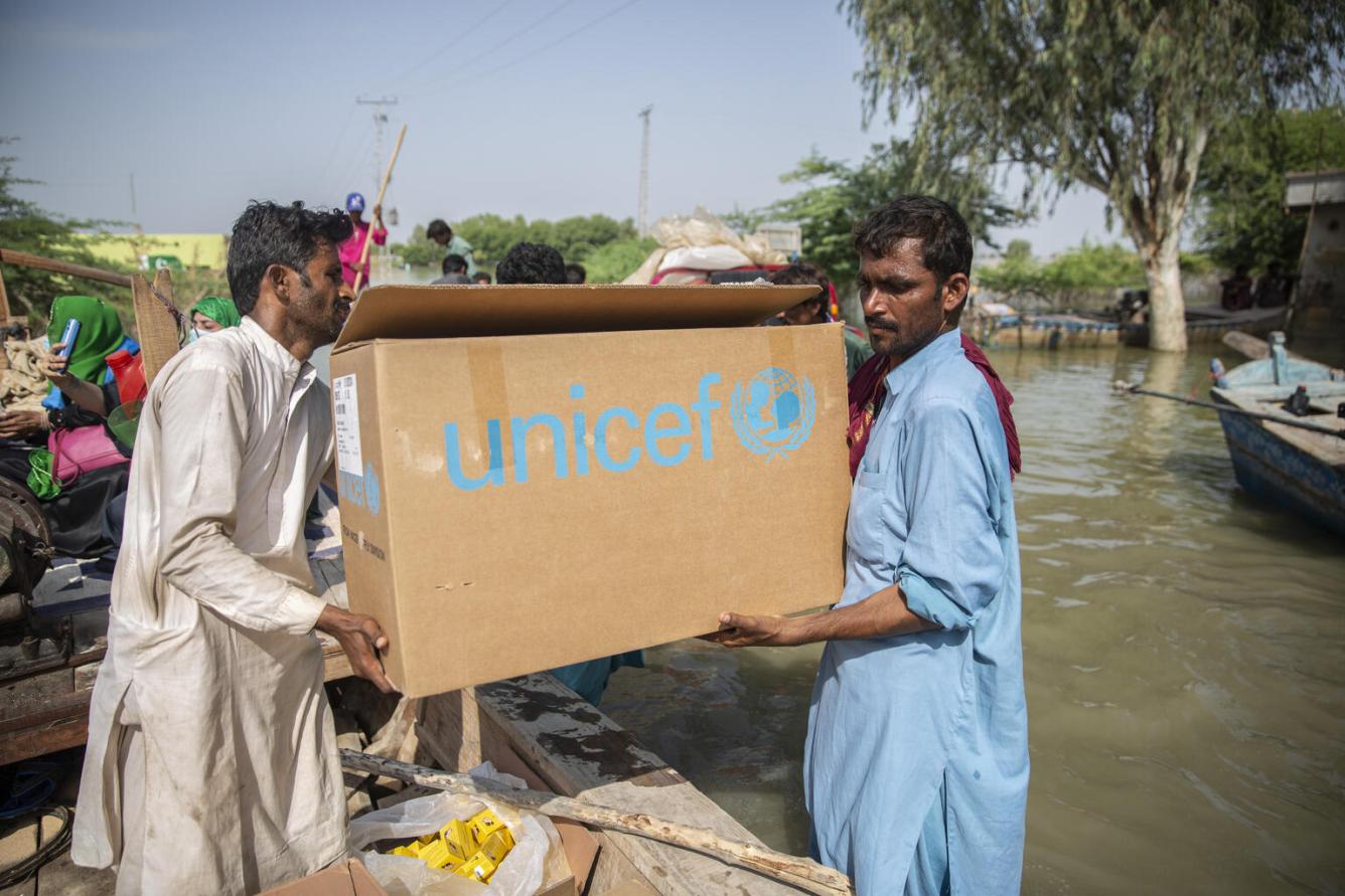 Deux hommes portent une boîte brune de l’UNICEF. On voit derrière eux les eaux de crue et un bateau.