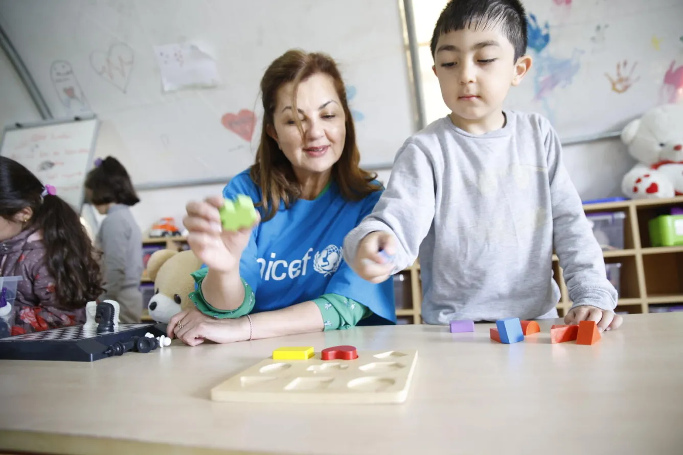 A woman sits at a table with a child who is standing up and showing some toys to her.