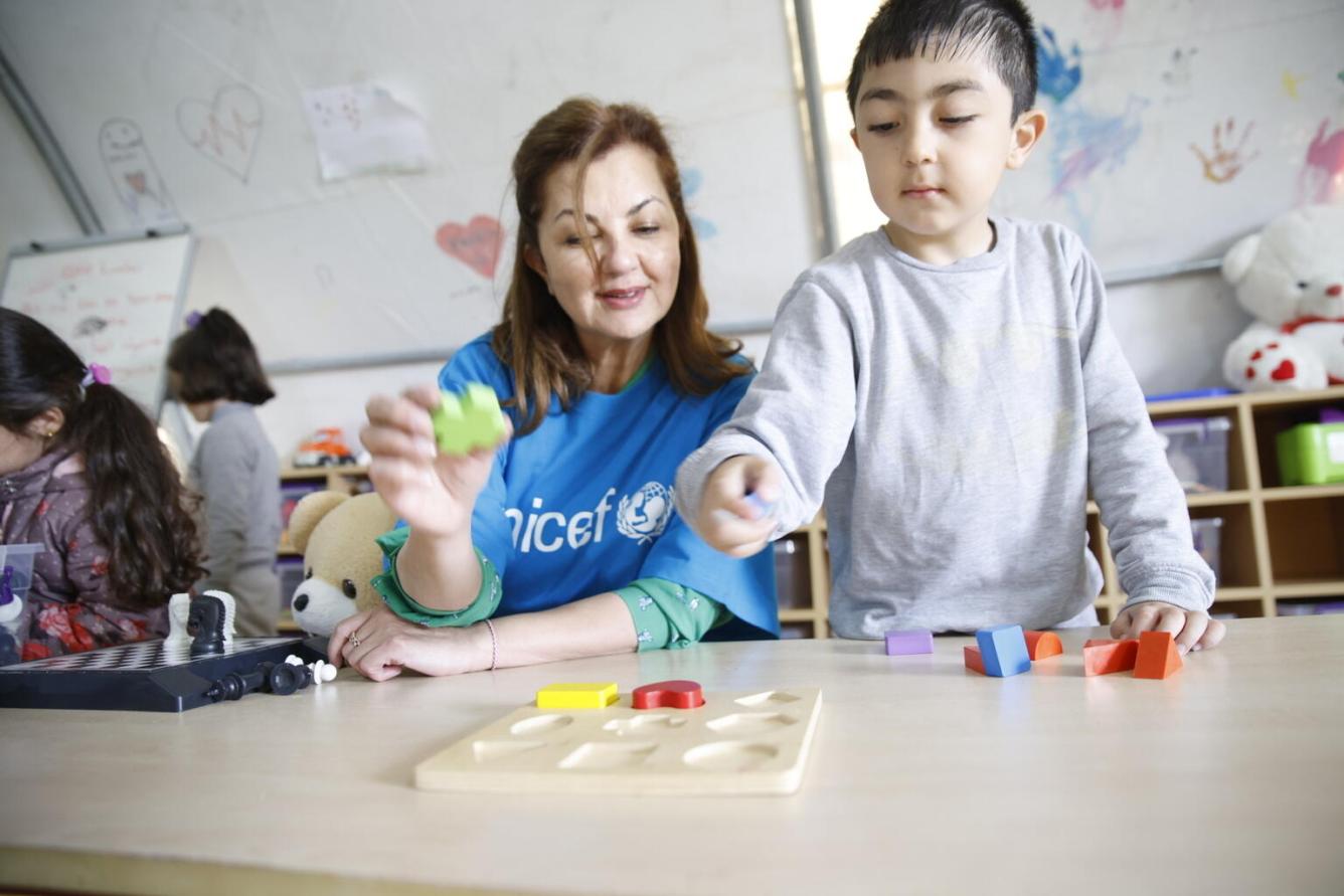 Une femme est assise à une table en compagnie d’un enfant, debout, qui lui montre des jouets.