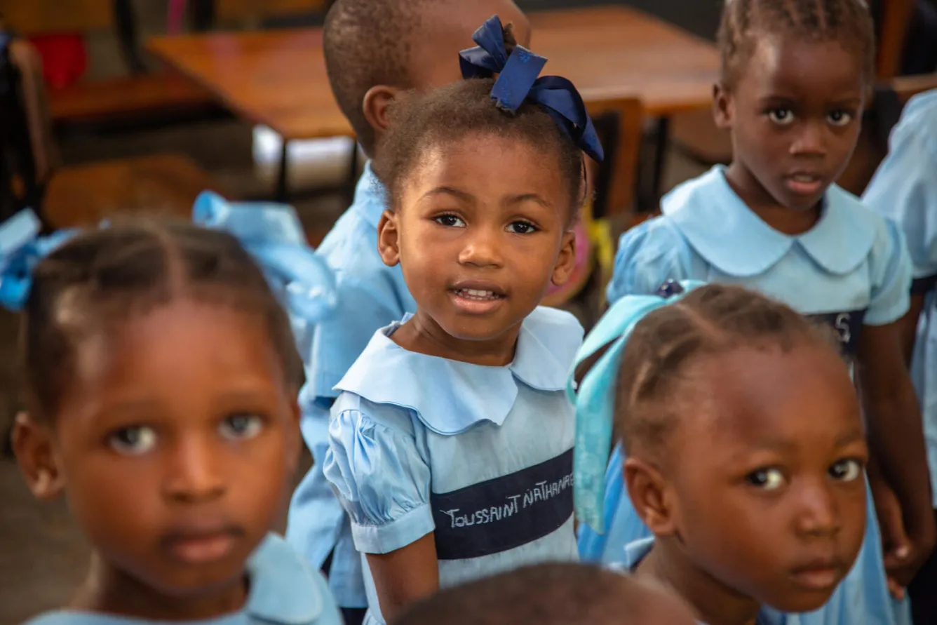 A group of children in blue uniforms sit on desks, a girl in the centre of the frame looks right at the camera with a slight smile.