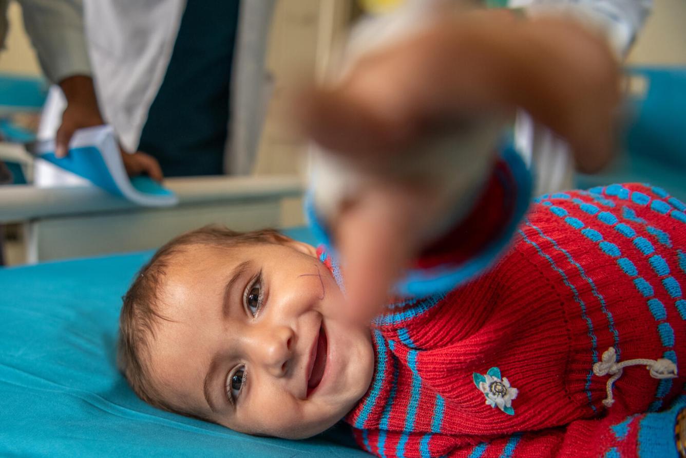 Un bébé allongé sur un lit tend le bras vers l’objectif en souriant.