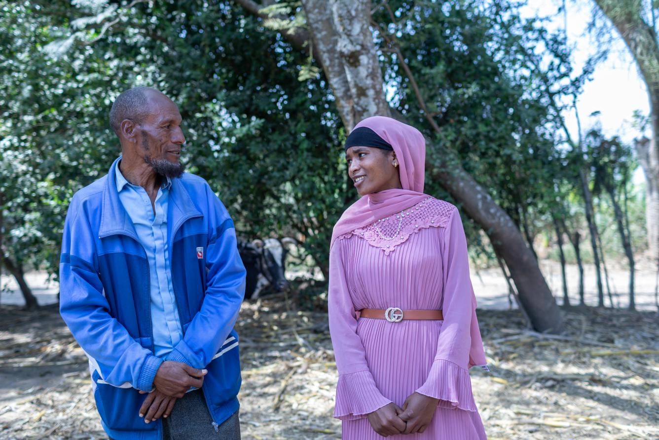 A man in dressed in blue and his daughter in pink stand in an outdoor setting, looking at each other as though in mid-conversation.