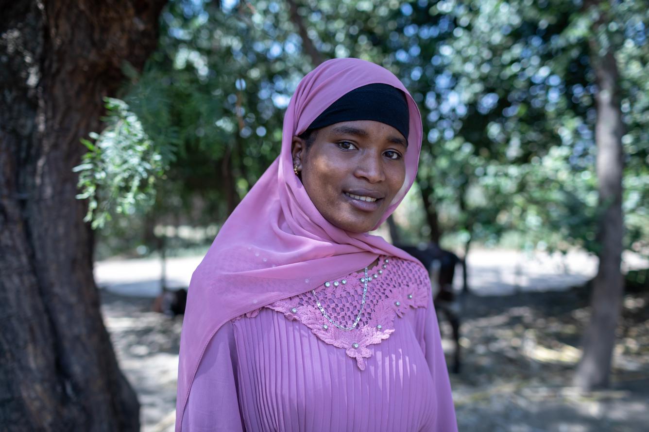 An adolescent girl in pink clothes looks directly at the camera with a slight smile. She is in an outdoor setting.