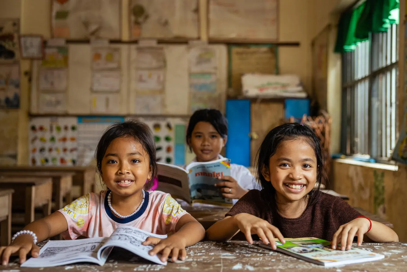 At a classroom in Cambodia, three girls sit at their desks with their workbooks open and smile at the camera.