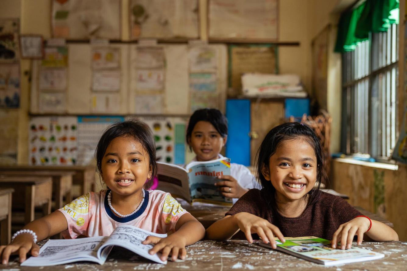 Dans une salle de classe au Cambodge, trois filles assises à leur pupitre avec leurs cahiers ouverts sourient à l’objectif.