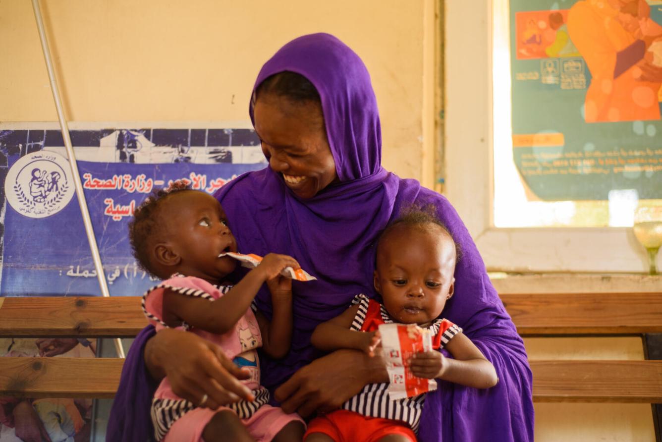 A mother holds her twin daughters who are eating from packets of ready-to-use-therapeutic food.