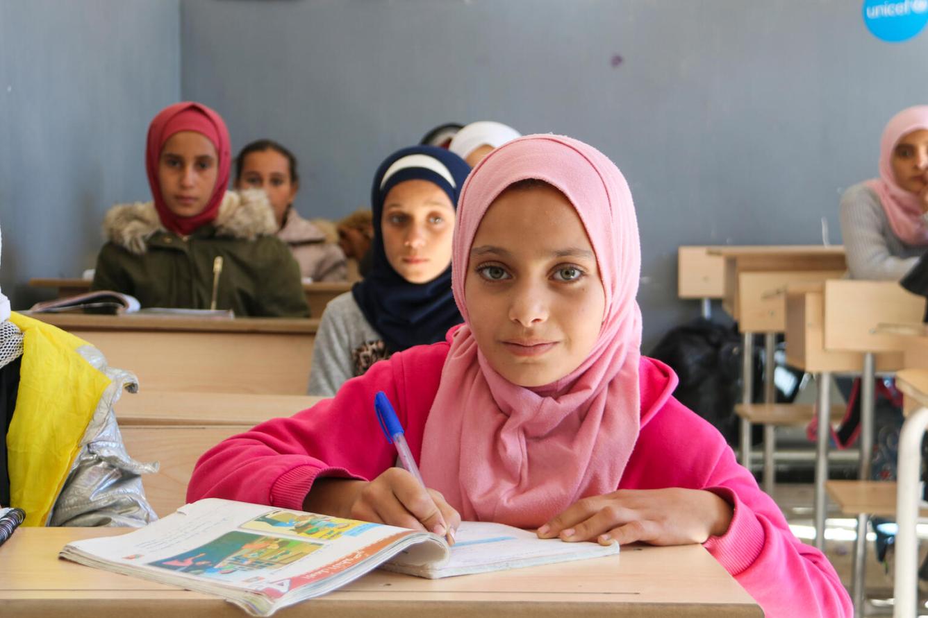 Dans une salle de classe en Syrie, une fille portant un foulard rose écrit dans son cahier et sourit à l’objectif.