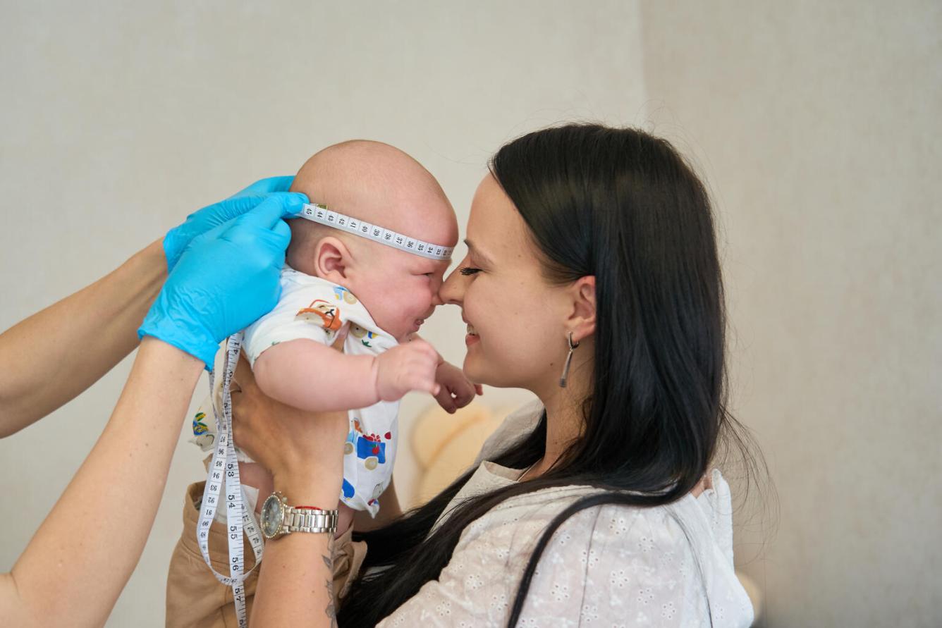 A nurse measure an infant's head with a measuring tape, as the child's mother holds the child close to her face with their noses touching. Only the nurses arms can be seen in the photo, with the mother and child in focus in center.