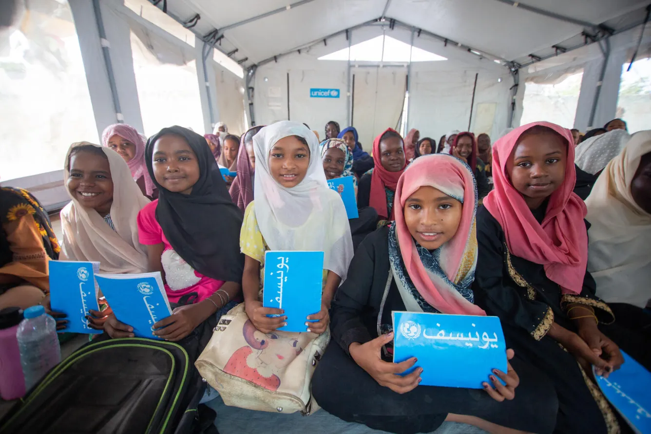 A group of children holding blue UNICEF notebooks smile at the camera at a UNICEF-supported safe learning space in Geziera state, Sudan.