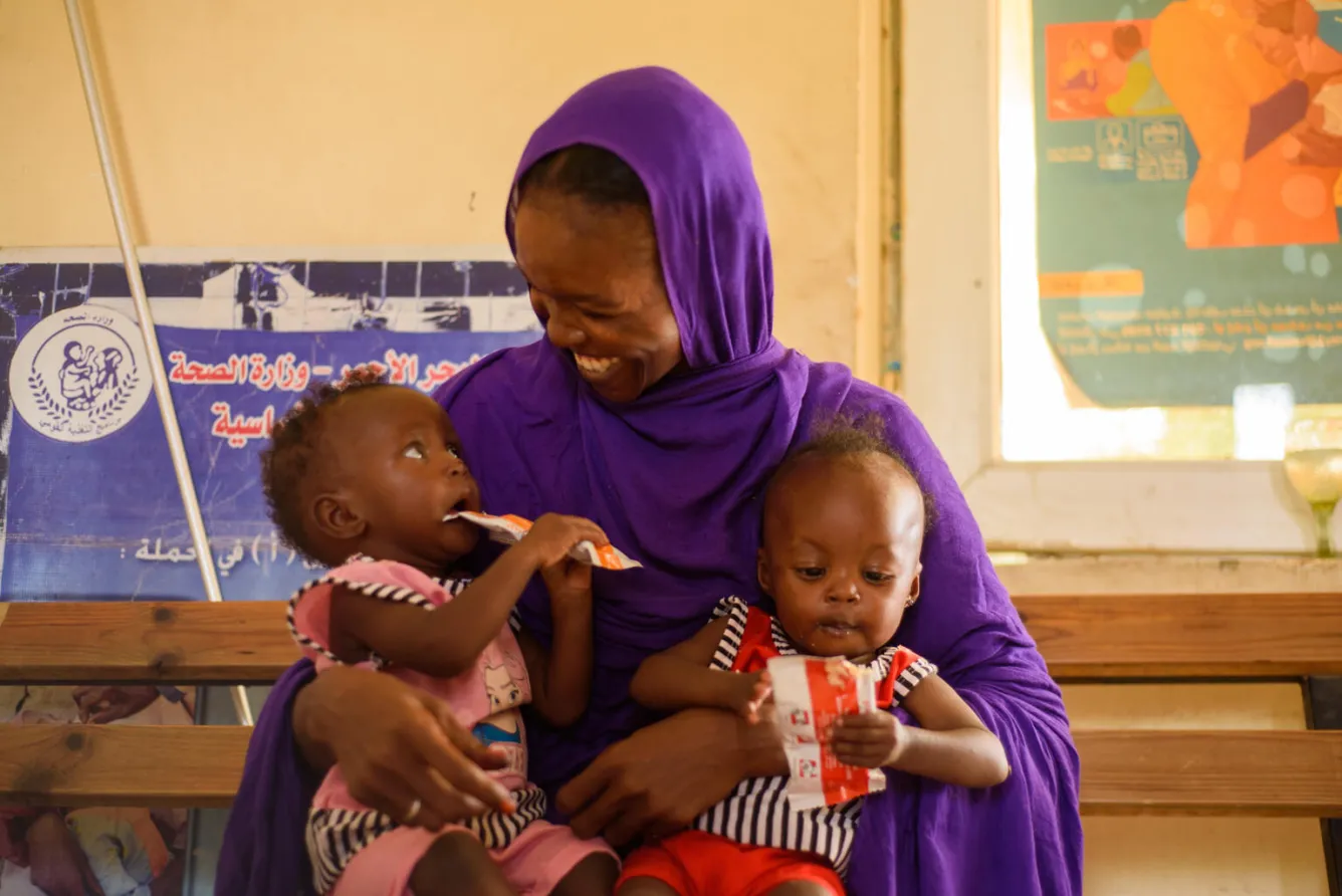 A mother in Sudan holds her two infants in her lap. Both children are holding RUTF sachets and the mother is looking down and smiling at the children.