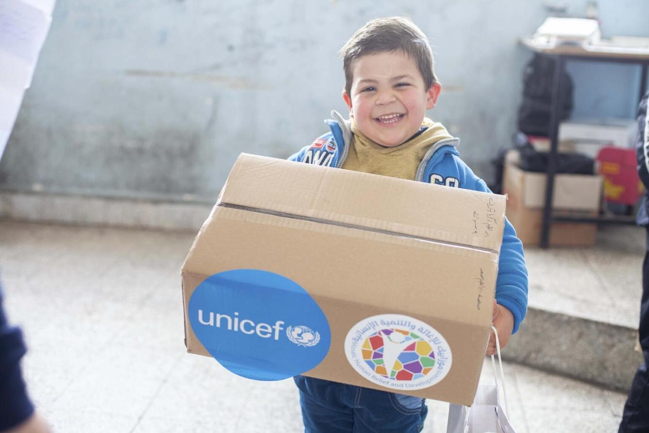 Eight-year-old Ali, smiles at the camera while holding a box of clothes he received from UNICEF at a collective shelter in Lattakia governorate, Syria.