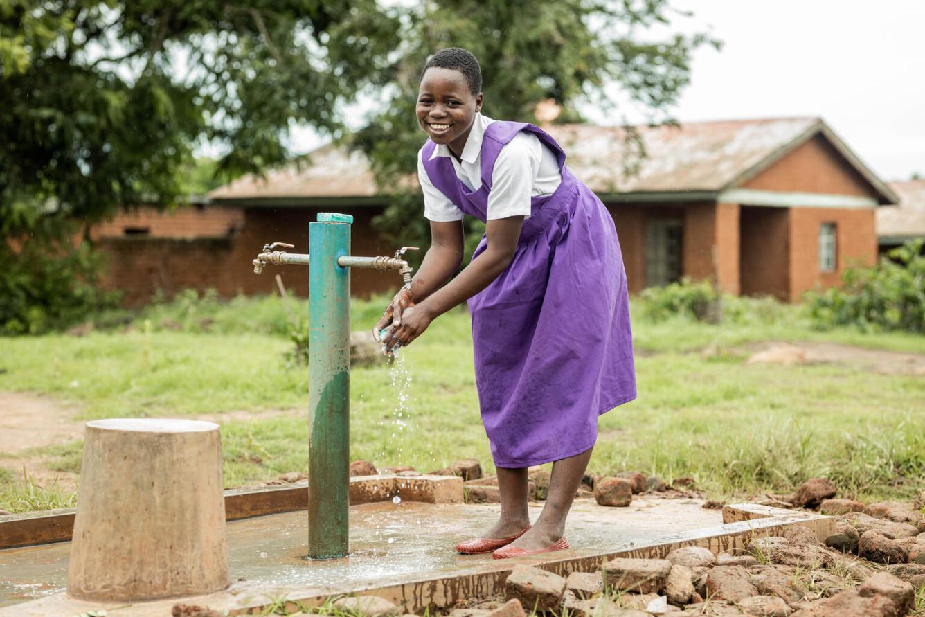 A girl smiles at the camera while washing her hands at an outdoor water pump.
