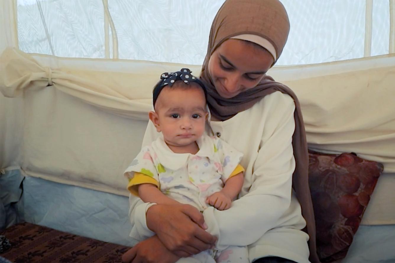 A child is seated and holding an infant on their lap inside a tented shelter.