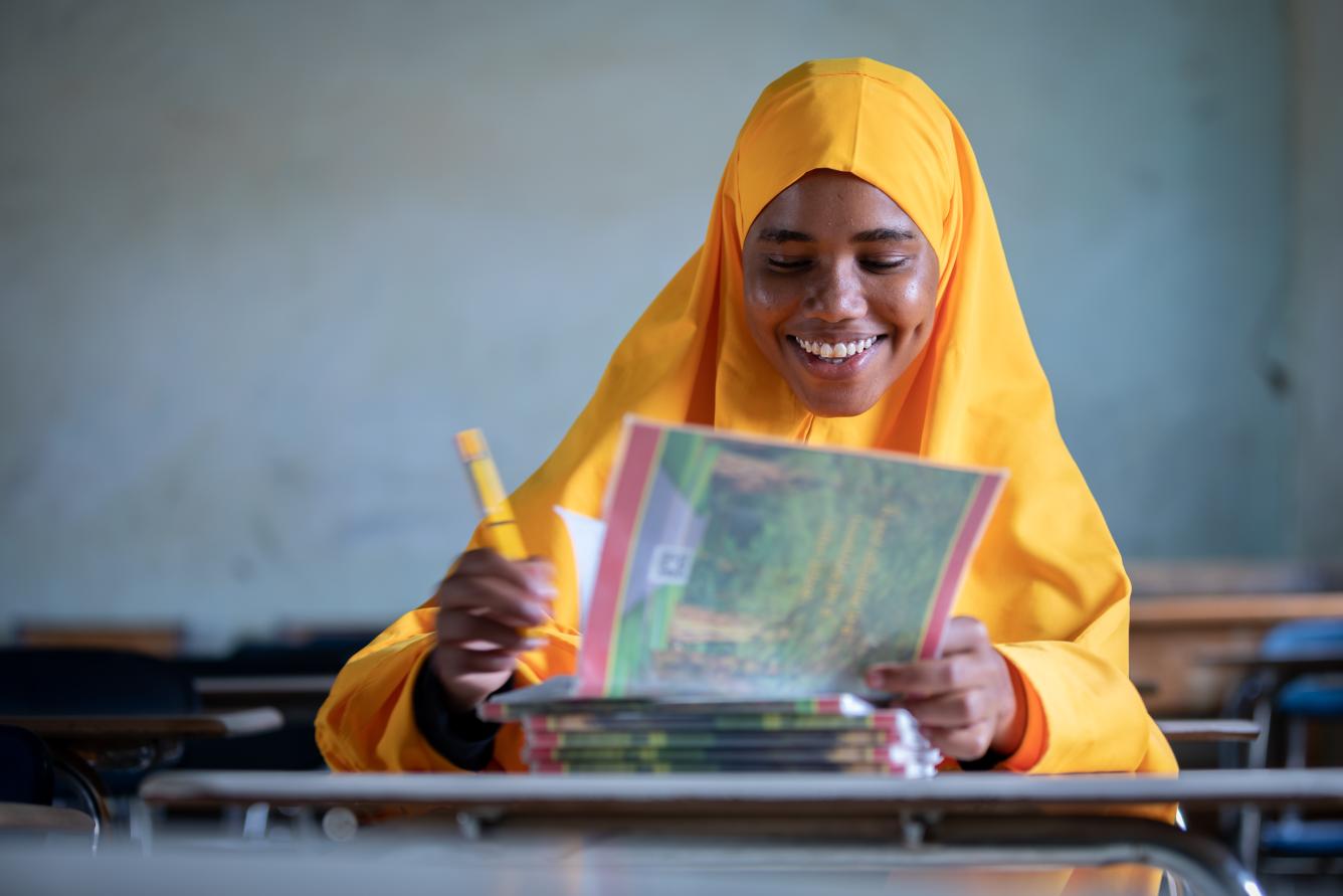 A girl, wearing tangerine clothes sits in her class in Somalia and looks towards the camera