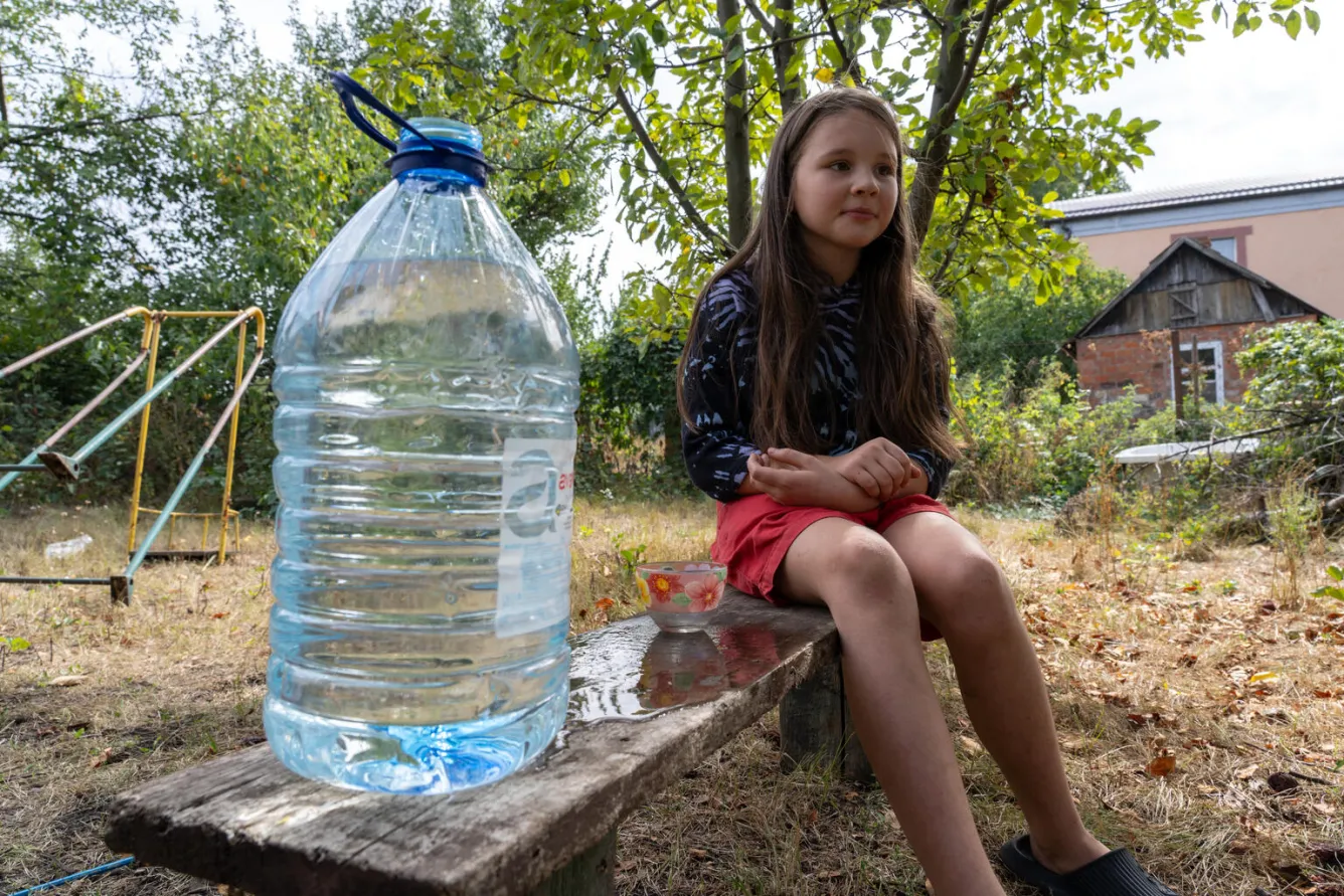 A girl sits outside on a bench with a large bottle of water beside her.
