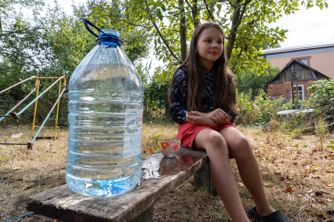 Une fillette est assise sur un banc à l’extérieur sur lequel se trouve également une grande bouteille d’eau.