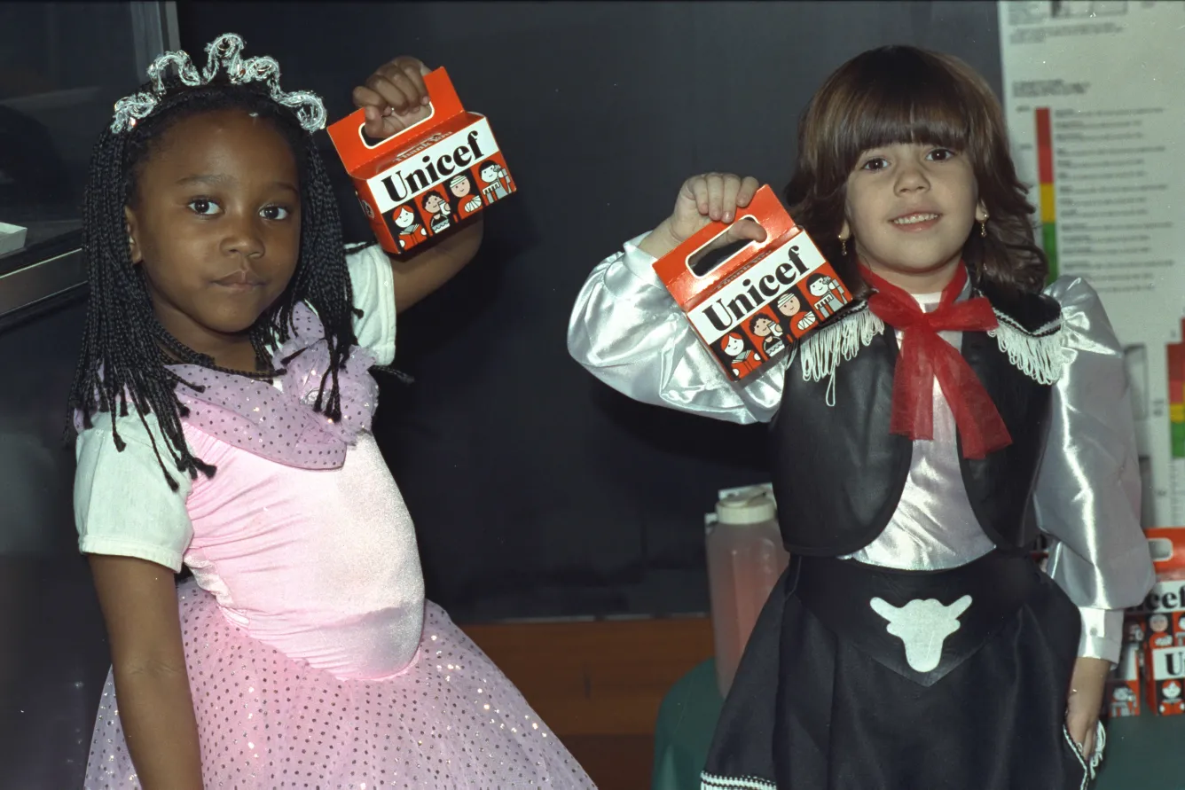 In a classroom, a child dressed up as a princess, and another dressed up as a cowboy hold up orange UNICEF Halloween boxes.