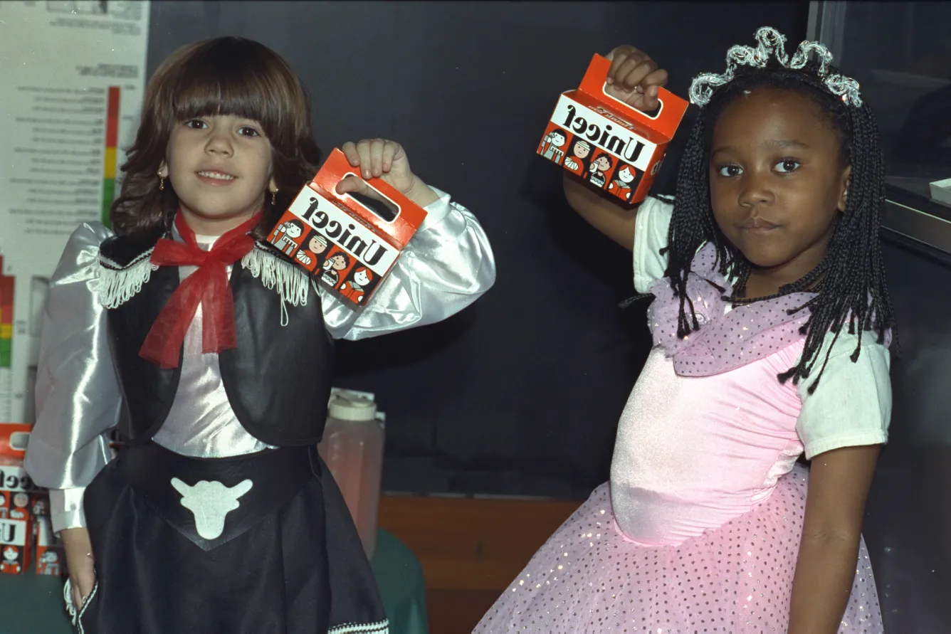 In a classroom, a child dressed up as a princess, and another dressed up as a cowboy hold up orange UNICEF Halloween boxes.