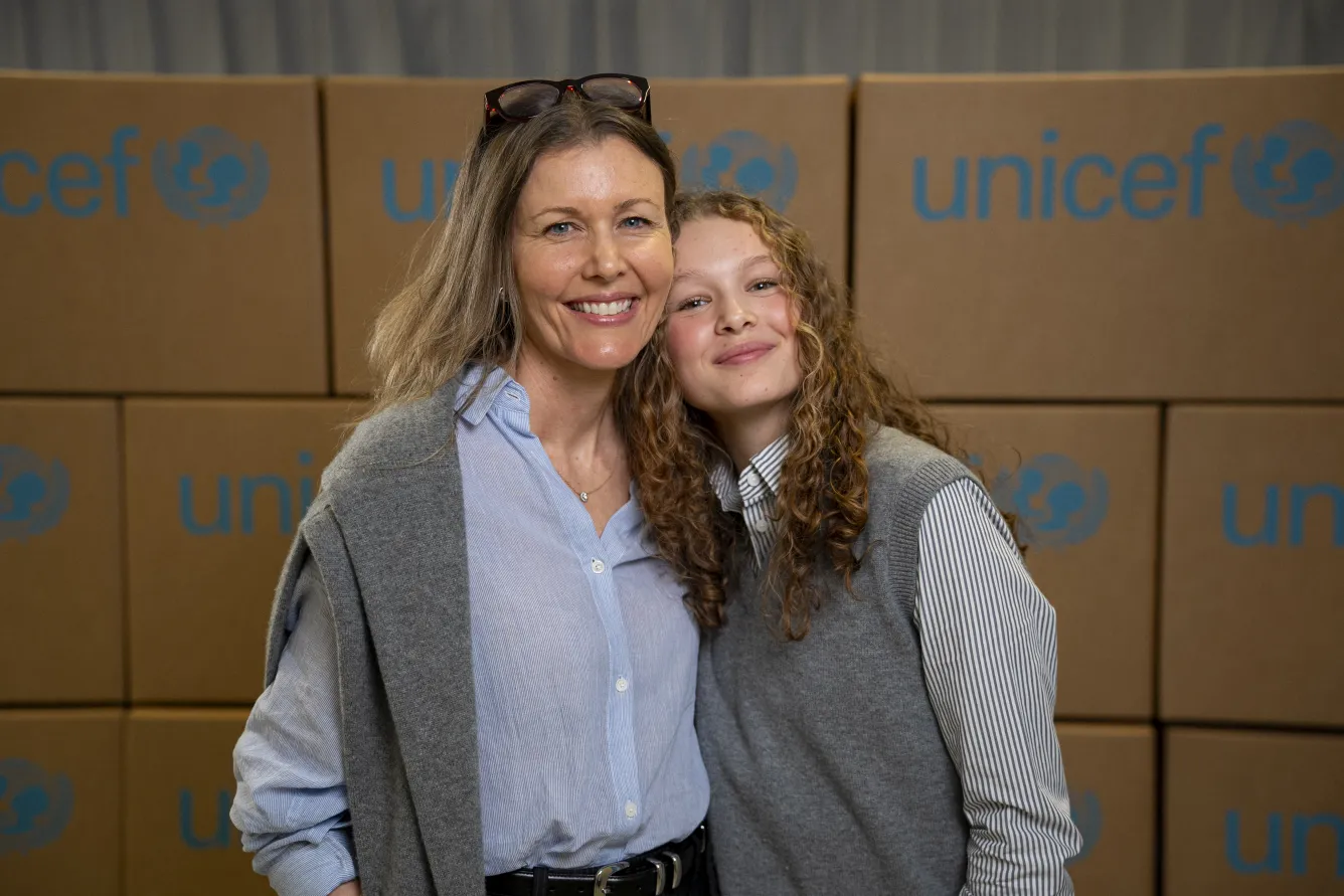 A woman and a young girl smile while looking at the camera. Behind them are cardboard boxes with the UNICEF logo on them. 