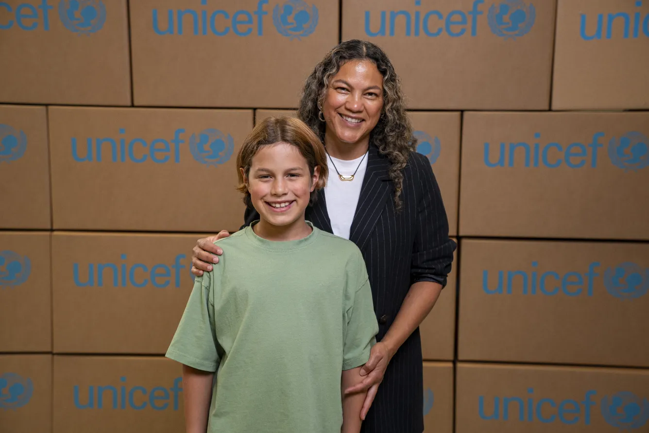 A woman and a young boy smile broadly, standing side by side in front of a backdrop displaying UNICEF cardboard boxes.