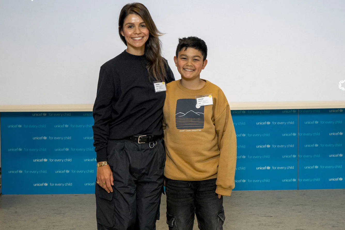 A woman and a young boy smile broadly, standing side by side in front of a backdrop displaying UNICEF's logo. The setting conveys a positive and friendly atmosphere.