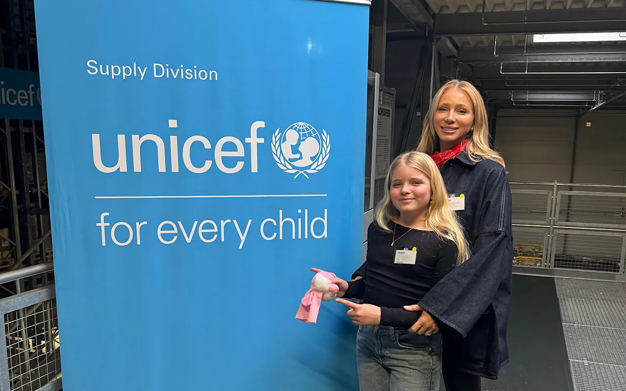 A woman and a young girl smile, standing in front of a UNICEF Supply Division banner, behind them is a warehouse setting.