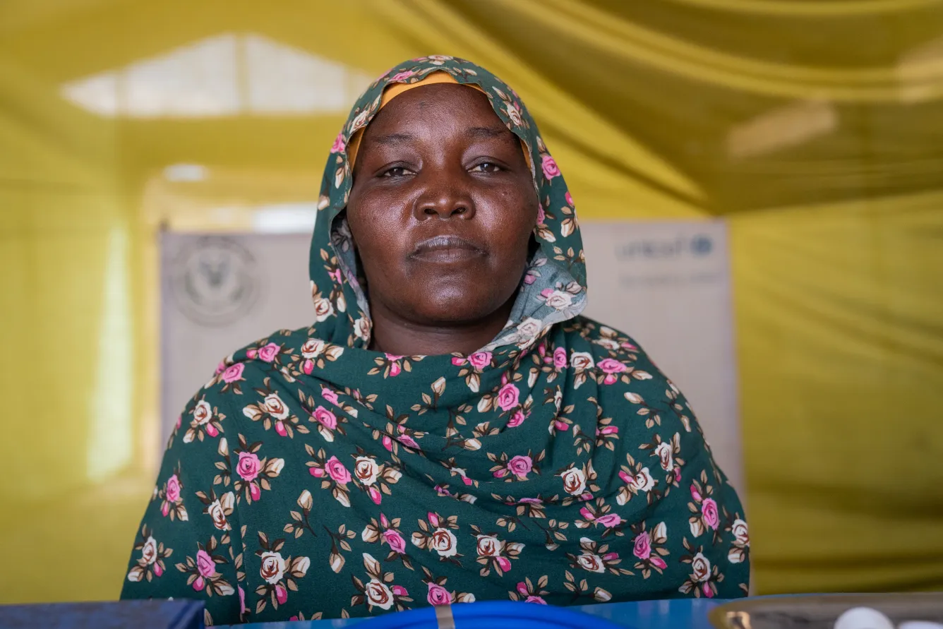  In a tent, a woman wearing a green headscarf looks at the camera. 