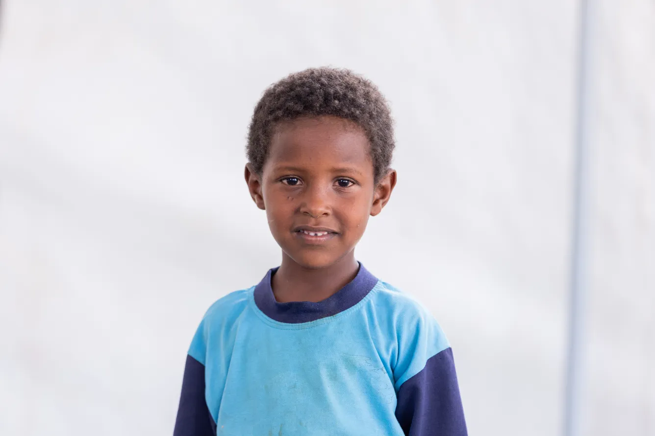  In a tent, a boy wearing a blue shirt smiles at the camera. 