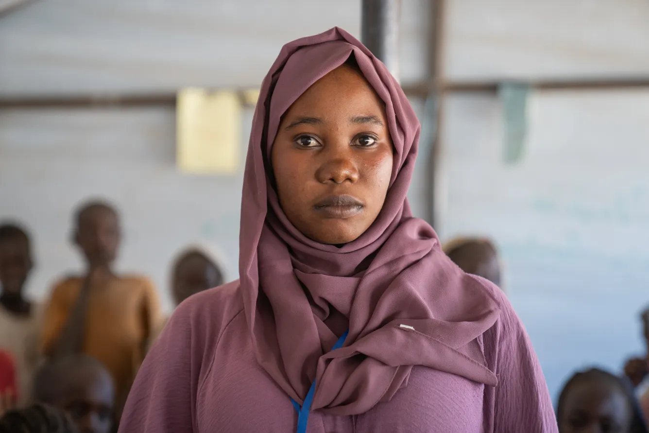  In a tent, a woman wearing a purple headscarf looks at the camera. 