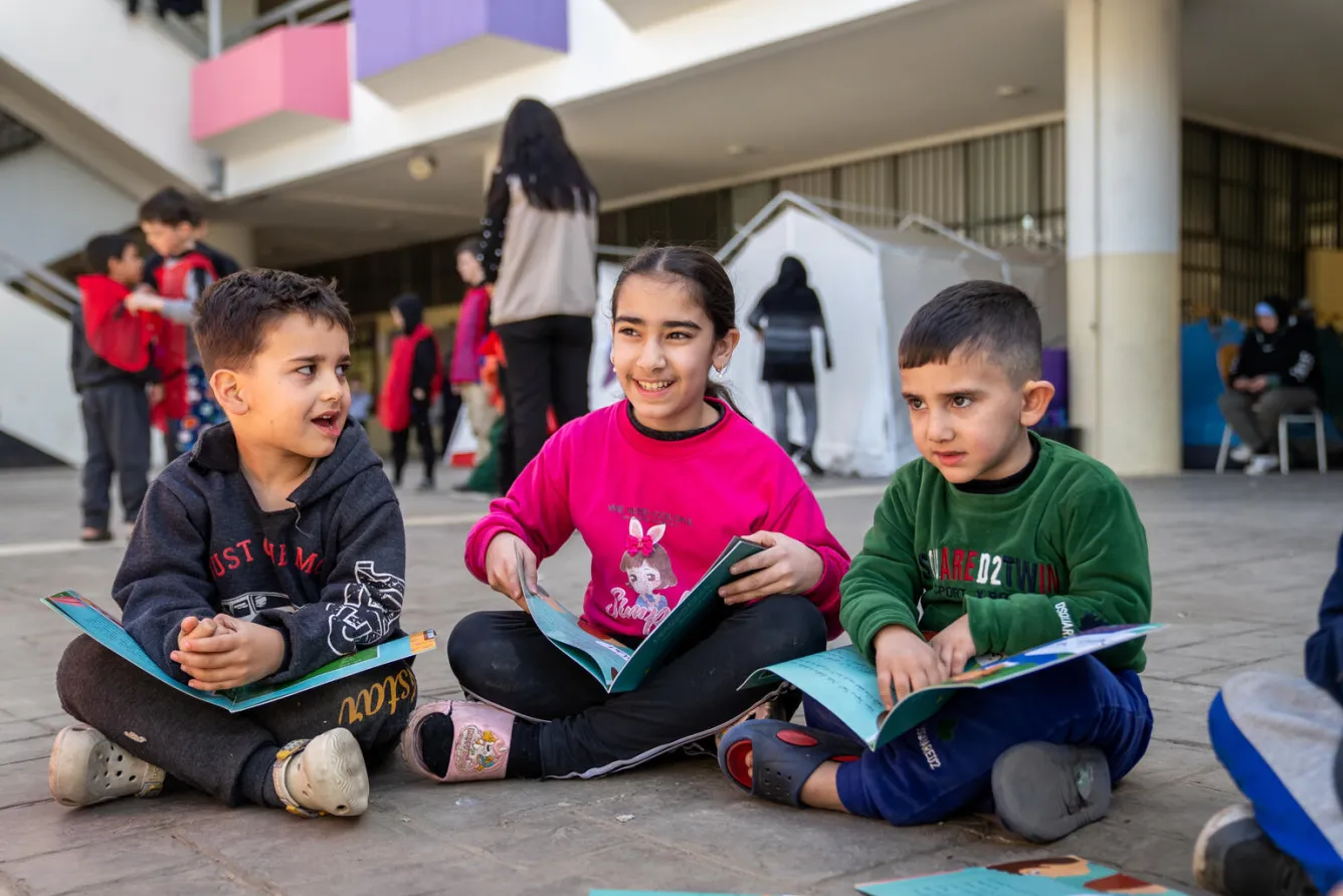 Three children sitting on the ground outdoors, each with an open book on their laps.