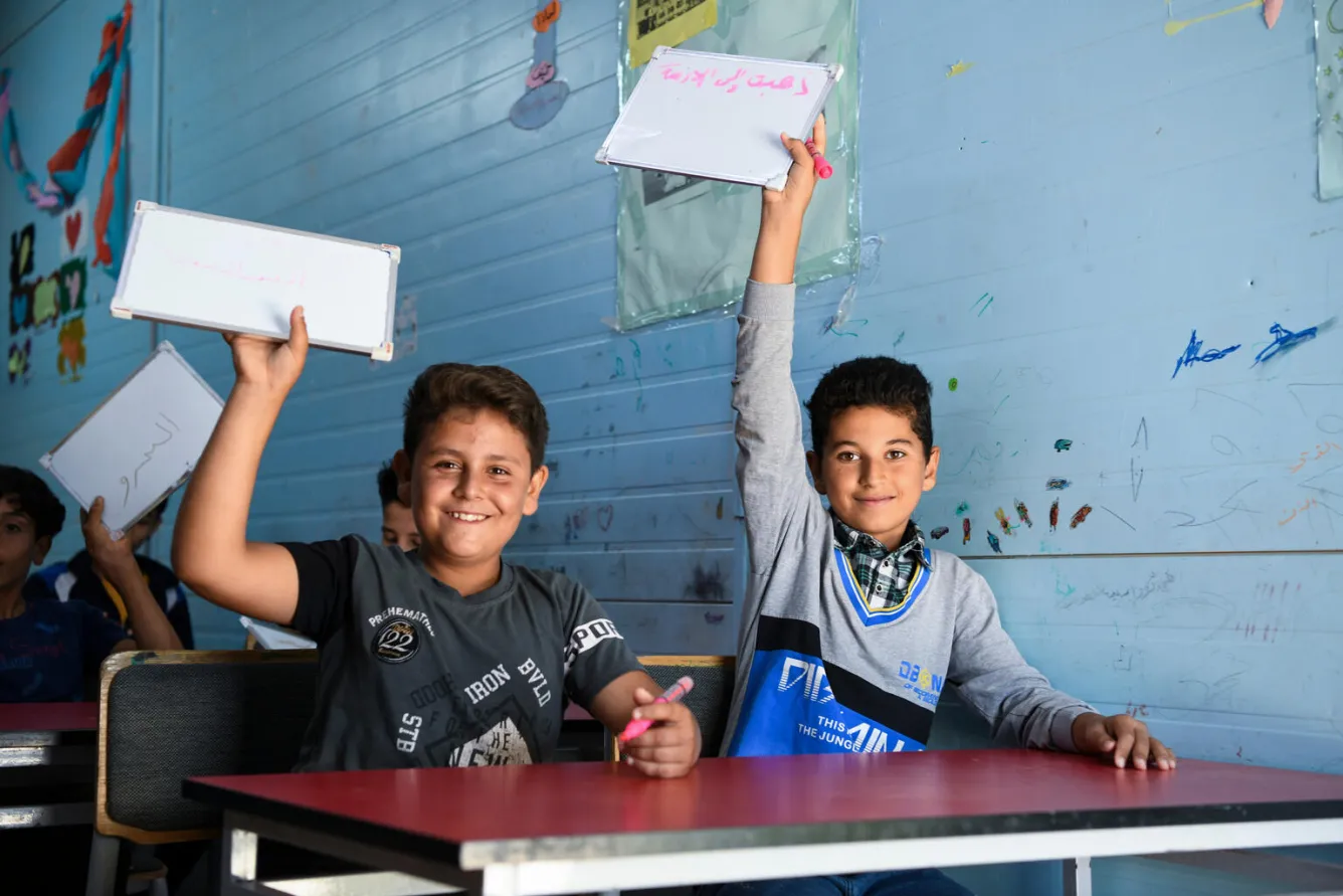 Deux garçons sont assis à leur pupitre dans une salle de classe et sourient à l’objectif tout en soulevant leurs petits tableaux blancs. 