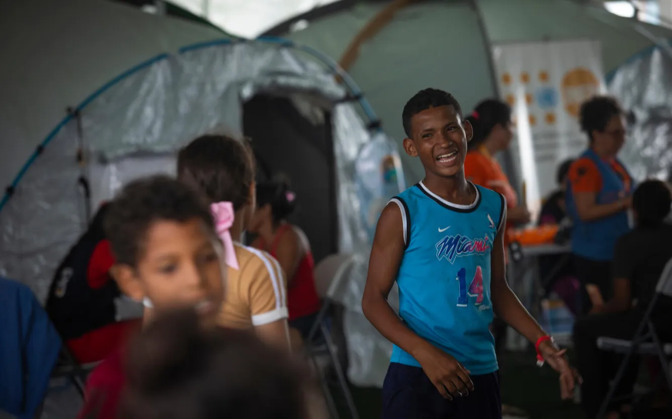 At a temporary camp, a boy wearing blue laughs among other children.  