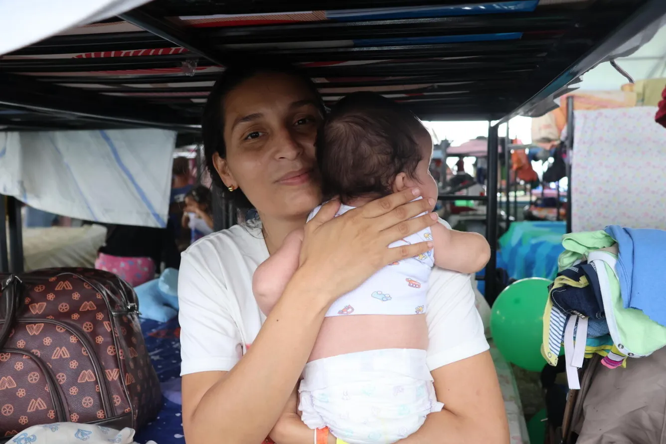 In a temporary camp, a woman holding a baby looks at the camera. 