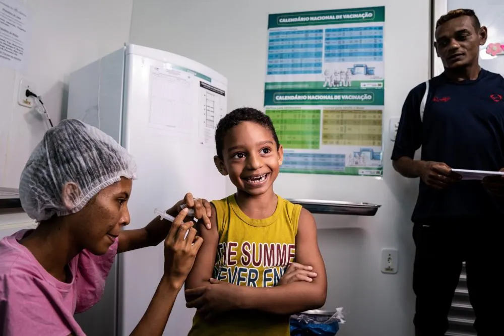 A child smiles as he receives a vaccination in his arm from a health worker in a clinic setting.