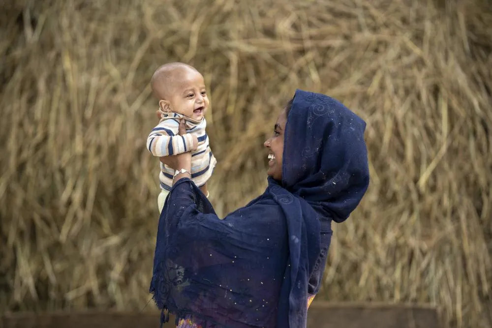 An infant laughs as he is playfully held up by his mother who is smiling back at him.
