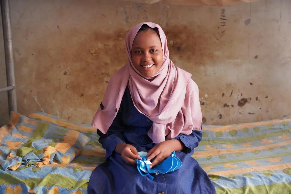 An adolescent girl sitting and smiling at the camera and holding sanitary towels.