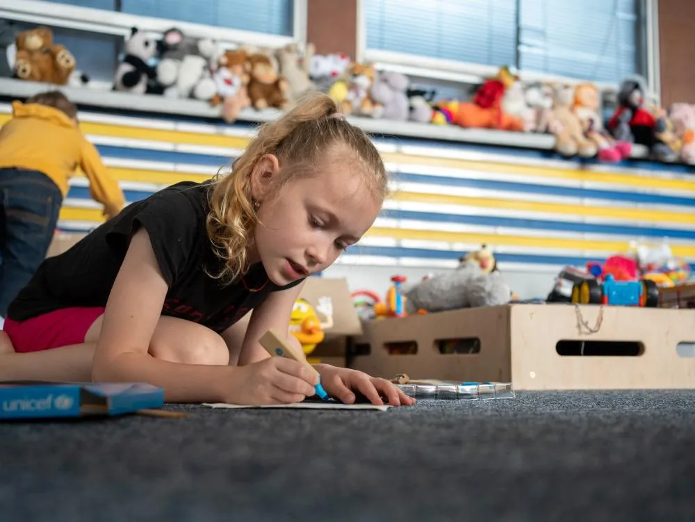 A girl sits on the floor drawing with a UNICEF activity kit inside a children’s centre.