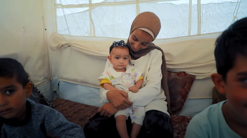 A child is seated and calmly holding an infant on their lap inside a tented shelter.