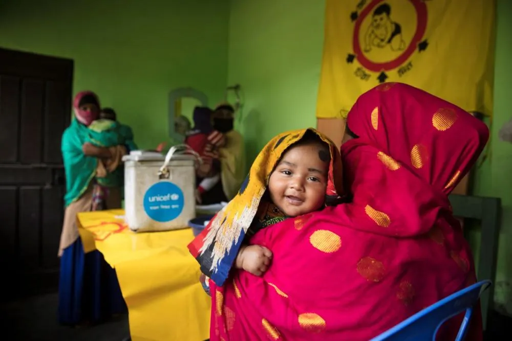 A toddler smiles at the camera over an adult’s shoulder in a health clinic setting.