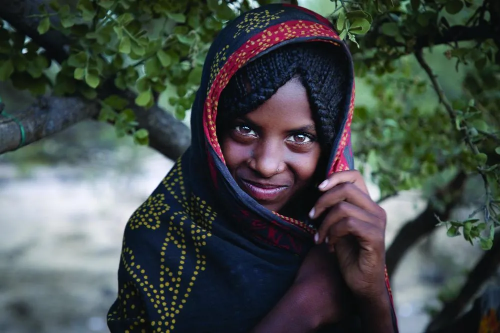 Portrait d'Aicha, onze ans, souriant à la caméra avec confiance en tenant le tissu d'un châle brodé sous son menton.