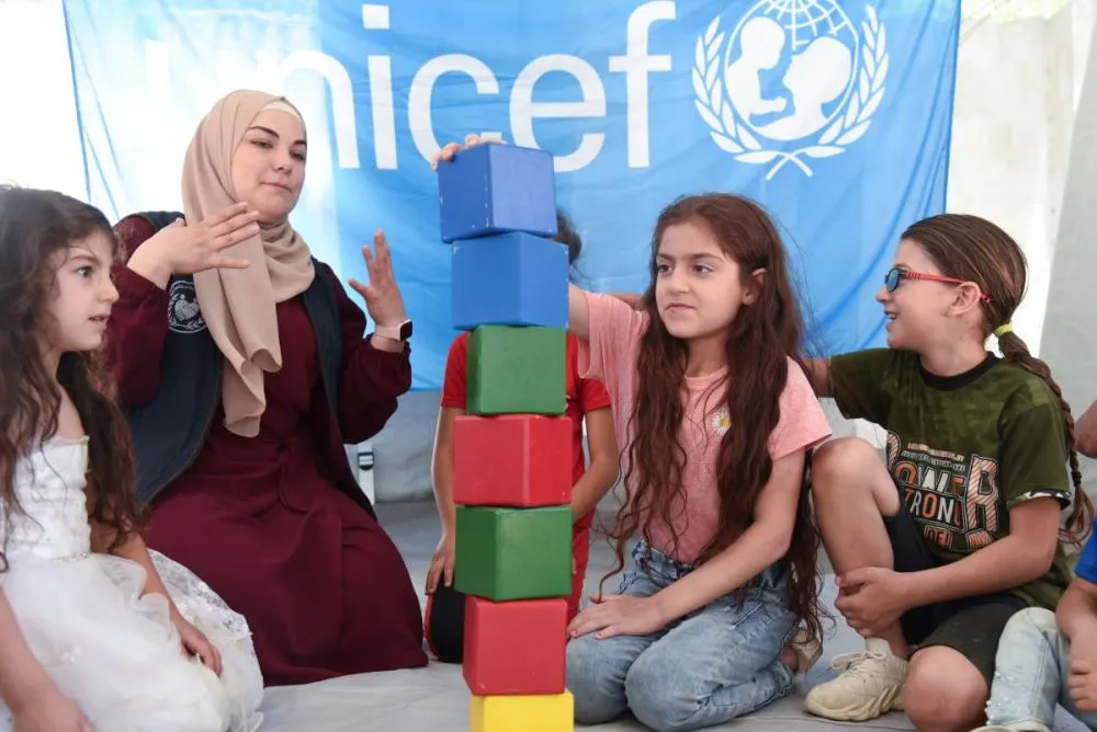 A child stacks large foam blocks on the floor as three other children and an adult watch, sitting in front of a wall displaying a UNICEF banner.