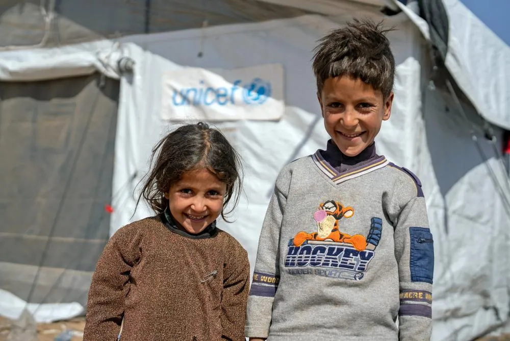Two children stand together smiling in front of a UNICEF tented shelter.