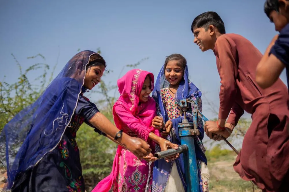 A group of happy children gathered around an outdoor water pump on a sunny day.