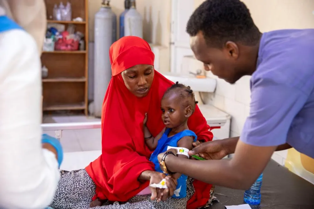 A child sitting with an adult in a health clinic setting having their arm circumference measured by a health worker.