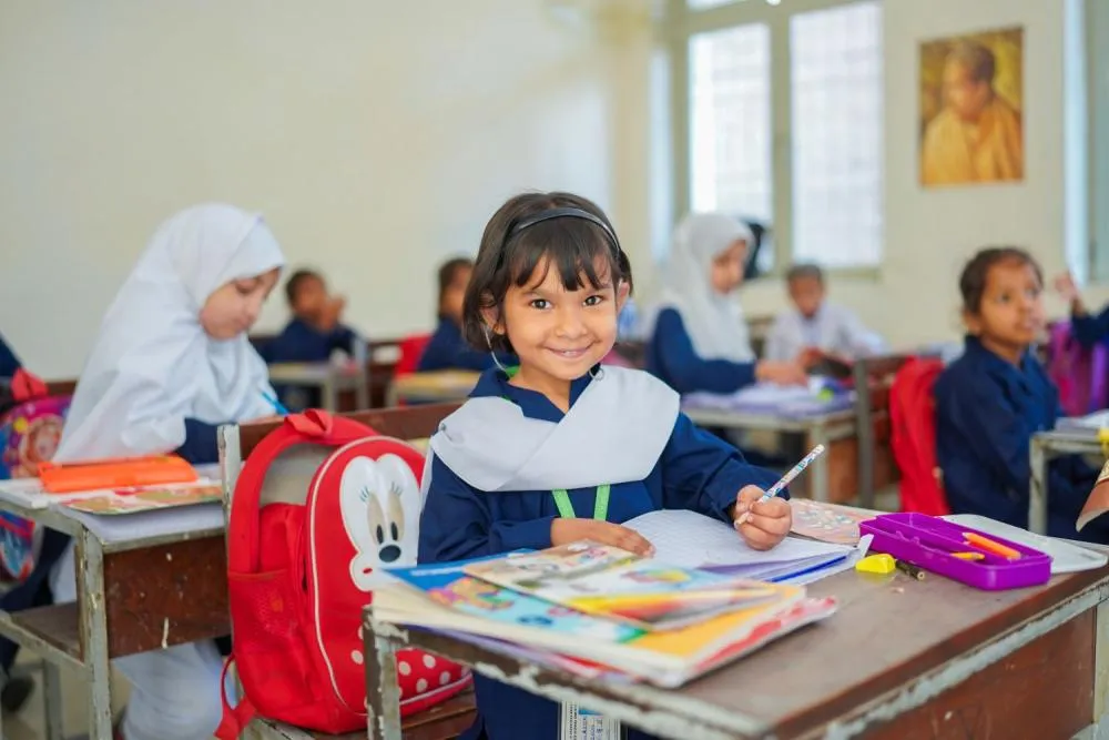 A young student sitting at a classroom desk holding a pencil over an exercise book and smiling at the camera.