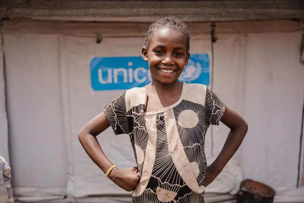 An adolescent girl stands smiling confidently with her hands on her hips in front of a UNICEF tented shelter.