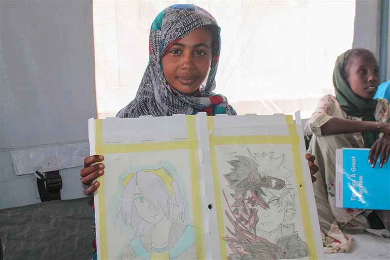 A girl wearing a headscarf holds up her drawings at the camera.